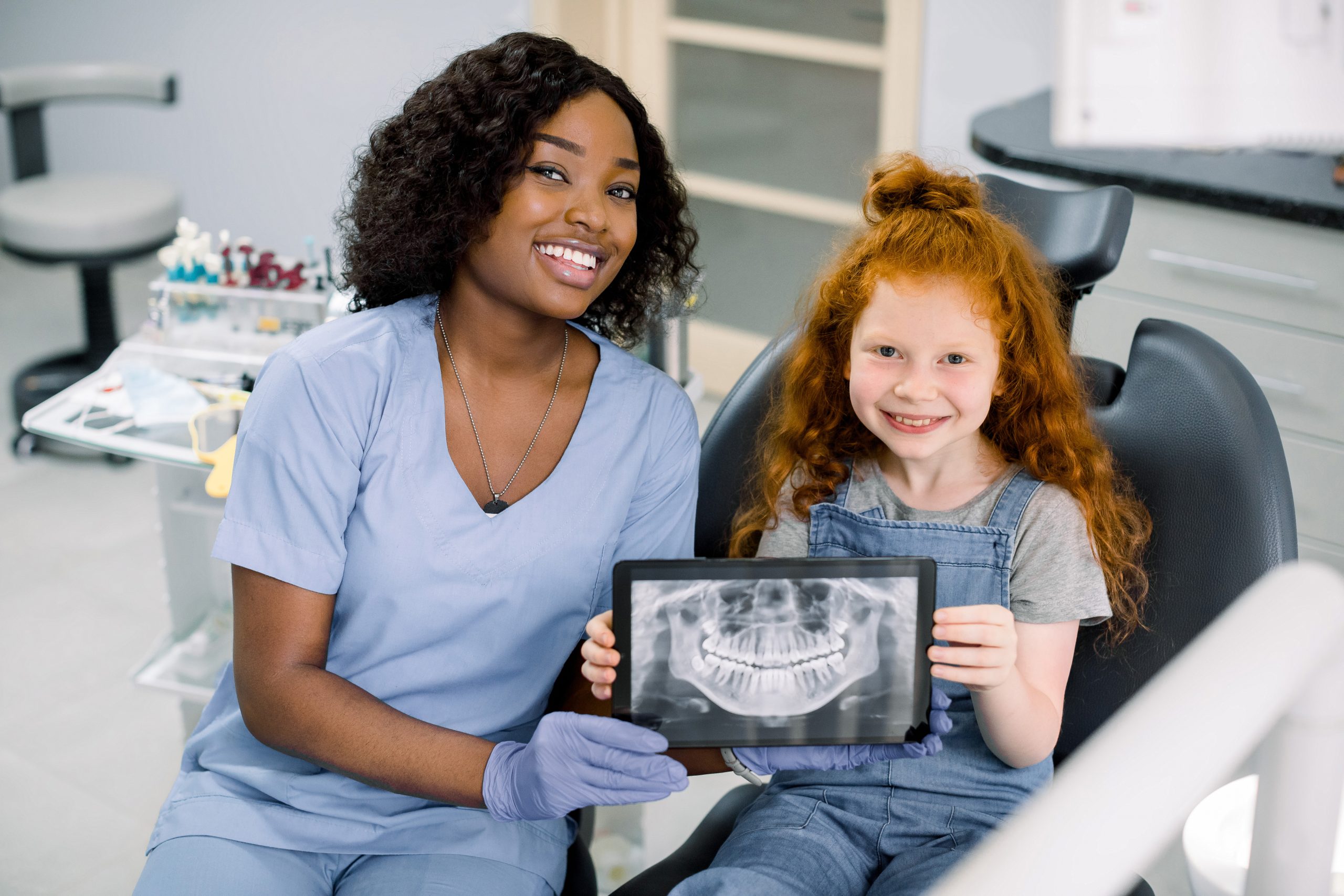 Little smiling girl with red curly hair sitting on chair and looking at camera, while holding x-ray scan image of her teeth on digital tablet together with her cheerful black female dentist at clinic.