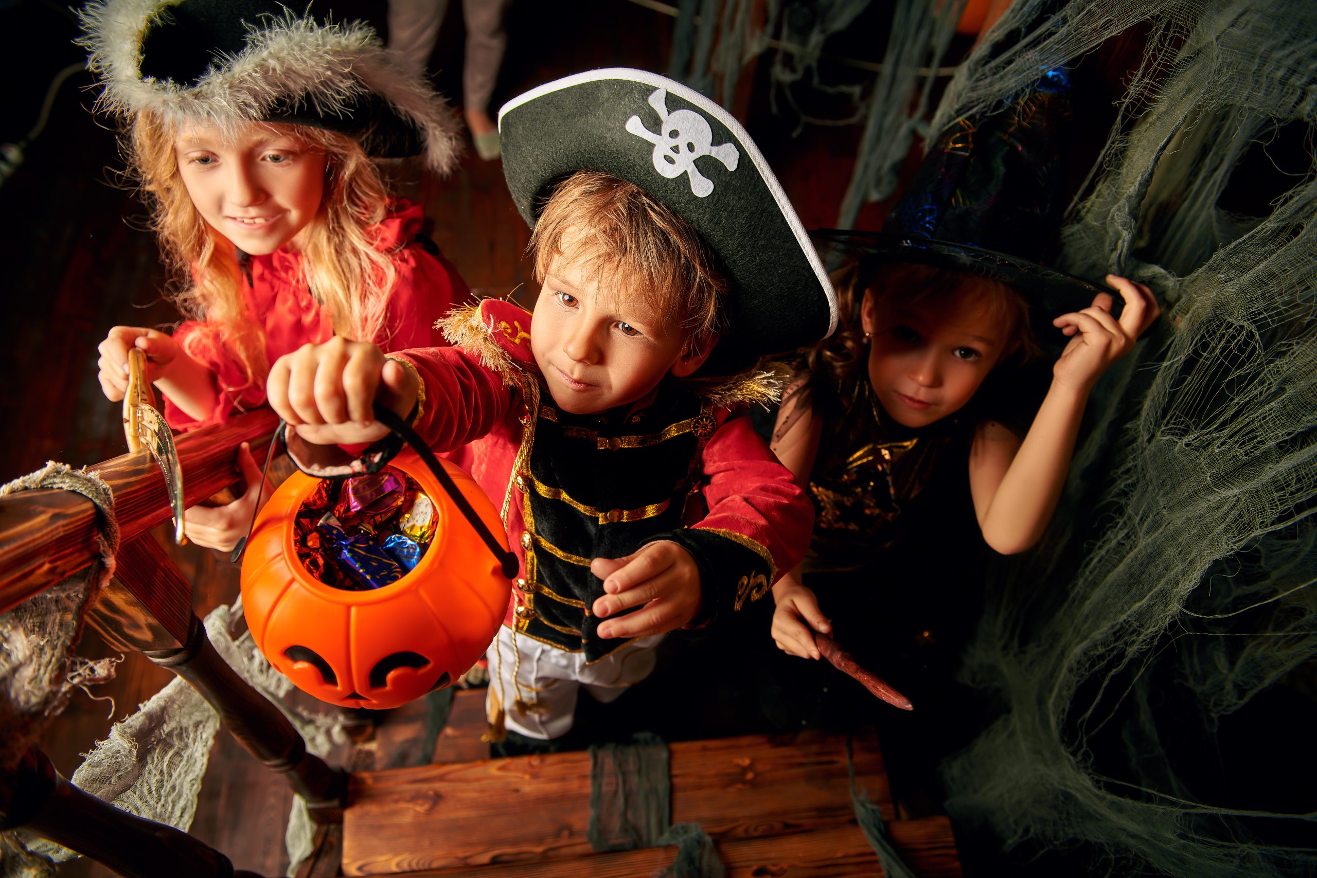 Happy children in carnival costumes trick or treating with pumpkin basket in the old wooden house. Halloween party.