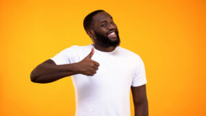 Young bearded black guy showing thumbs up, smiling on camera yellow background